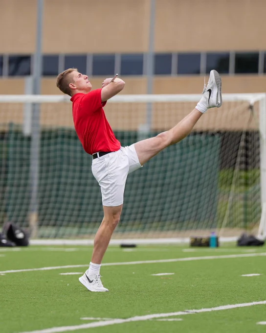 Ben Fox is strutting with a soccer goal in the background. He is wearing a red shirt, white shorts, and athletic shoes.