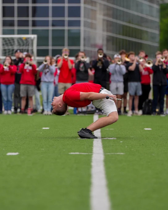 Jacob who is in a red shirt and white shorts performs a dramatic backbend. In the background, a marching band plays instruments. 