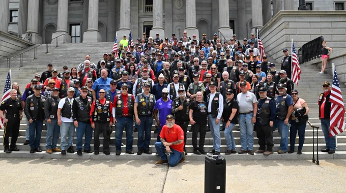 A large group of people, many wearing motorcycle gear and vests, are gathered on the steps of a grand building with tall columns. The group is positioned in front of multiple American flags.