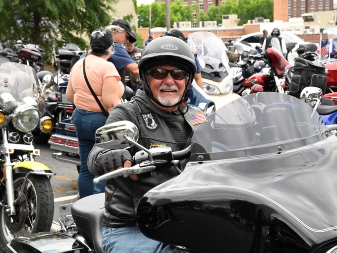 A person sits on a Harley-Davidson motorcycle in a parking area filled with other parked motorcycles. They wear a helmet, sunglasses, and a black leather vest with an American flag patch. People and various bikes are visible in the background.