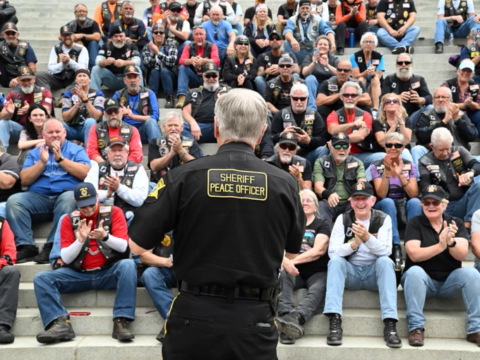 A person wearing a "Sheriff Peace Officer" uniform stands facing a large group of people seated on steps. The group is diverse, dressed in casual attire, many wearing vests with patches. Some are clapping, and there are numerous motorcycles in the background.