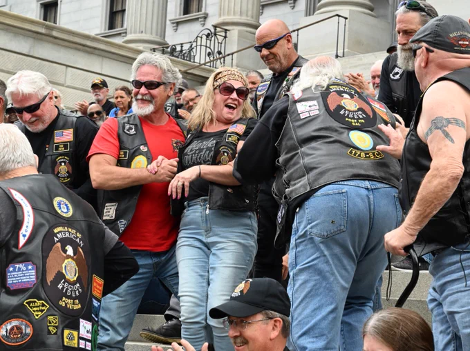 A group of motorcyclists wearing leather vests with various patches stands together on outdoor steps. They appear to be socializing and smiling. Some vests display emblems including "America's Guardians" and other badges. The setting is in front of a building with large columns.