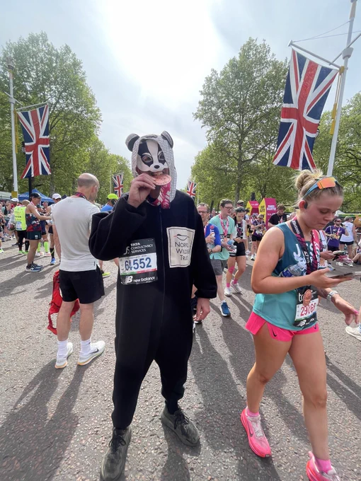 Patrick Barkham in a badger onesie and badger face-mask standing on the Mall biting his medal at the colourful finish-line of the 2026 London Marathon.