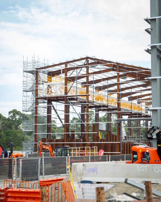 Construction site featuring a large steel framework for an acoustic shed, surrounded by scaffolding. Several orange excavators and construction barriers are visible in the foreground. Trees and a partly cloudy sky are in the background.