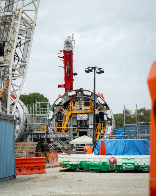 A large tunnel boring machine is positioned on a construction site, surrounded by scaffolding and equipment. The scene includes bright orange barriers, blue tarps, and a prominent red vertical structure in the background. Construction materials and cranes are also visible.