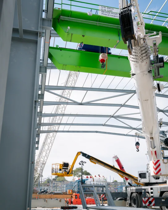 A construction site with large cranes and industrial machinery. Steel framework and beams are visible in the partially constructed building.