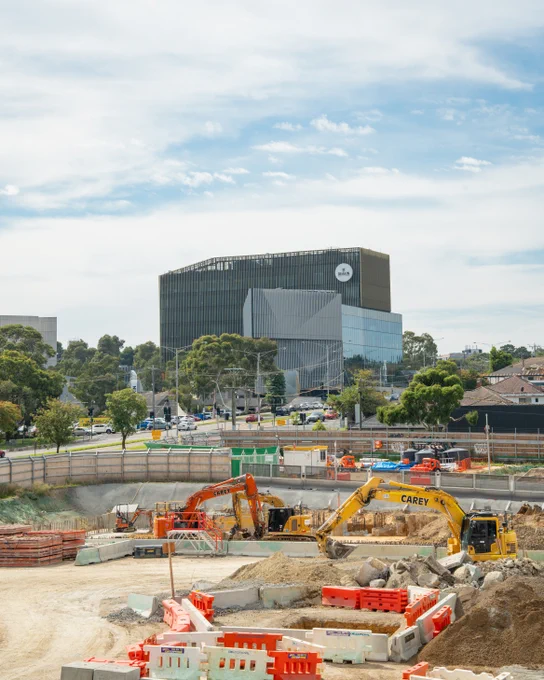 Suburban Rail Loop construction site with heavy machinery, surrounded by safety barriers. In the background, Deakin University Burwood campus is visible, with trees and roads nearby.