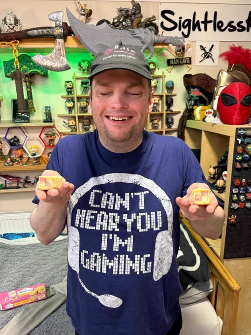 SK stands smiling and wearing an RNIB Design For Every Gamer cap, as well as a shirt with the words "Can't hear you, I'm Gaming" on it. He's holding two mini Battenbergs, the signature cake of the Game Accessibility Conference, one in each hand, positioned below and to either side of his face.