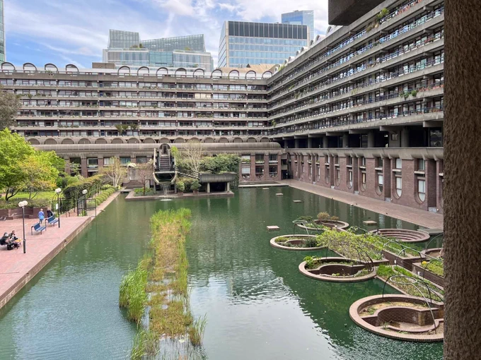 A urban courtyard with a pond in the foreground and large building at the back with balconies. Moss in the middle of the pond. Benches and trees line the left side with people walking and sitting down.