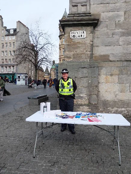 A police officer in uniform and high-visibility vest stands at a table with leaflets in Hunter Square.
