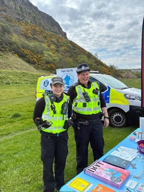 Two smiling police officers in high-visibility vests stand in front of a police van at an outdoor event, with a hill in the background.