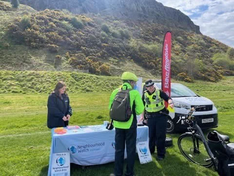 Community event with Neighbourhood Watch and police promoting bike safety outdoors, with a hill in the background.