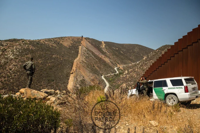 Two Border Patrol Agents scanning the U.S./Mexico border with binoculars in the mountains near the border wall under a clear blue sky.
