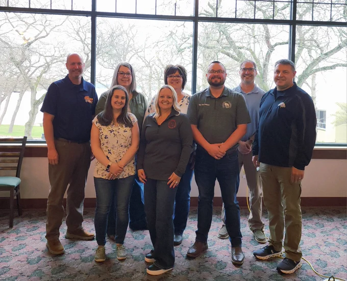 Group photo of Representative Alex Dallman (third from the right in a dark gray polo shirt) and seven other Green Lake County elected officials and staff standing indoors in front of large windows. They are at the Green Lake Conference Center after the Arbor Day Breakfast.