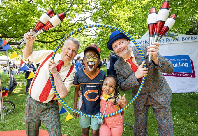 Two clowns and two children with face paint pose with juggling pins and a hula hoop at an outdoor event.