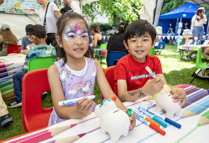 Two children decorating piggy banks at an outdoor craft table, one with face paint and the other with a temporary tattoo.