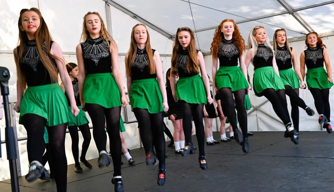 Young women in black tops and green skirts performing Irish dance on a stage.