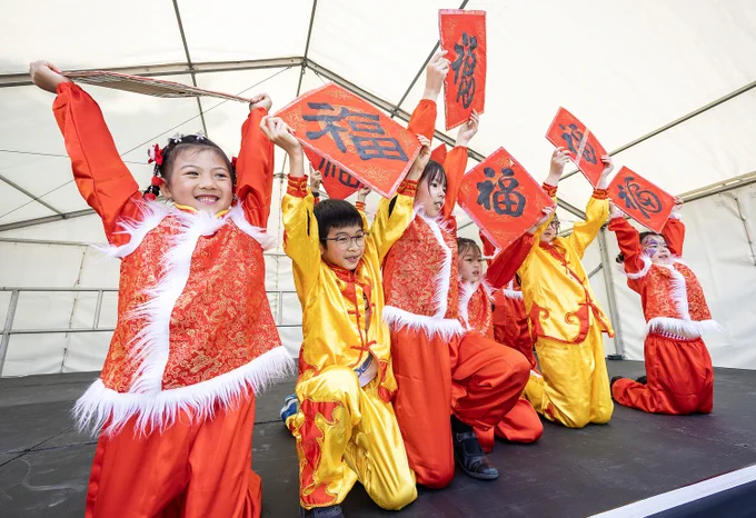 Children in traditional Chinese costumes perform on stage, holding red scrolls with calligraphy.