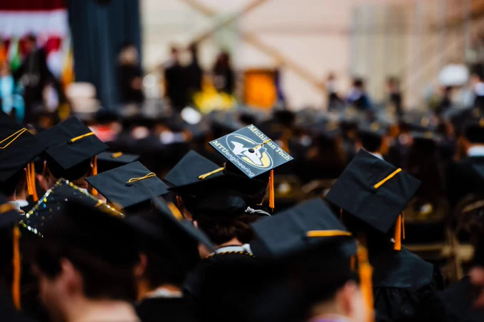 Michigan Tech graduates wearing black caps and gowns seated during a commencement ceremony, with one cap decorated with a cartoon character and text.