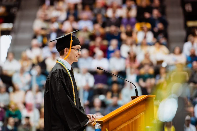 Michigan Tech graduate in cap and gown delivering a speech at a podium during a crowded commencement ceremony.