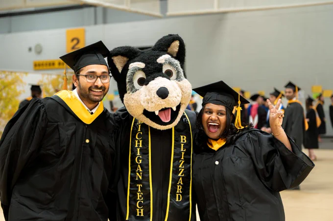 Two graduates in black caps and gowns pose with Michigan Tech's Blizzard mascot wearing a black and gold stole indoors.