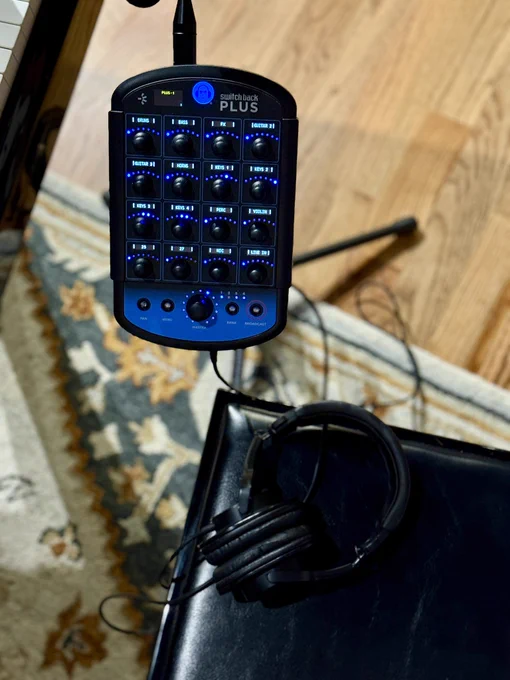 A Switch Back PLUS mixer   interface (a tablet-sized device with channel knobs and illuminated label screens) is mounted on a mic stand in a recording studio. A set of headphones is connected to it.