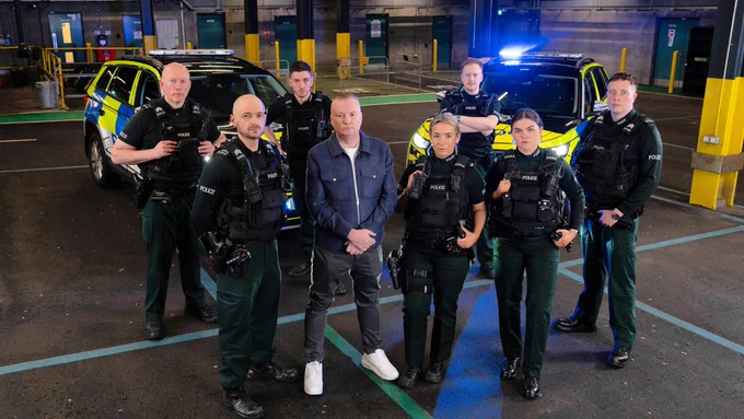 Seven police officers standing with Stephen Nolan with two police vehicles in the background.