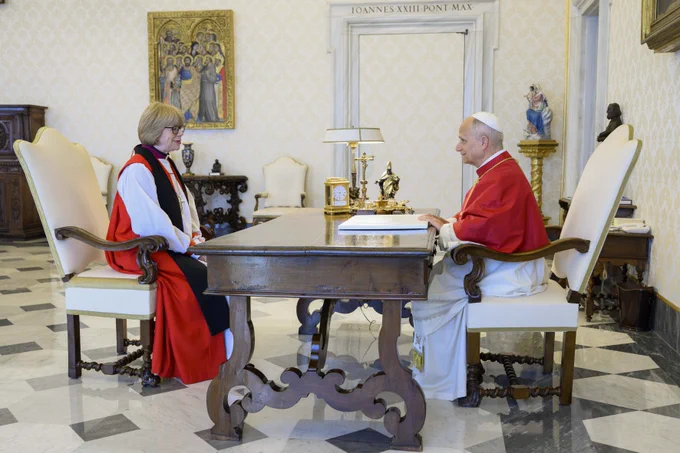 Pope Leo XIV people sit across from Archbishop Sarah at a large wooden desk in a Apostolic Palace. Pope Leo XIV wears white robes with a red cloak, while Archbishop Sarah wears black, white and red robes. The room features ornate decor, including a painting and a statue.
