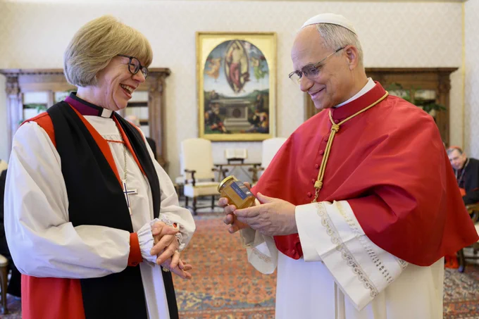 Pope Leo XIV receives a gift of Lambeth Palace honey from Archbishop Sarah. Pope Leo XIV is smiling and looking at the honey. Archbishop Sarah is smiling.