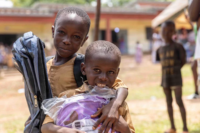 Two young Ivorian boys stand side-by-side at the Ouffoué-Diékro Primary School, smiling as they hold up brand-new, colorful backpacks. The boys are outdoors at their school during a donation ceremony held as part of Exercise Flintlock 26.