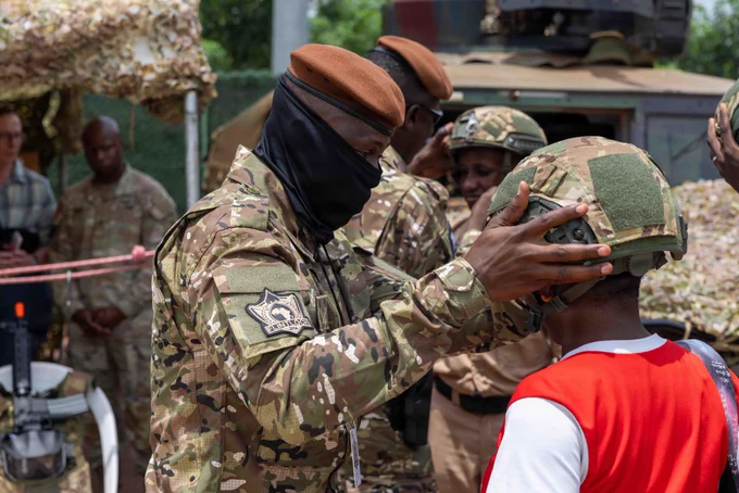 An Ivorian Special Forces soldier in tactical gear gently places a large military ballistic helmet onto the head of a young student. The student is smiling during a public service career day at the Lycée Scientifique de Yamoussoukro, with other students watching the interaction.