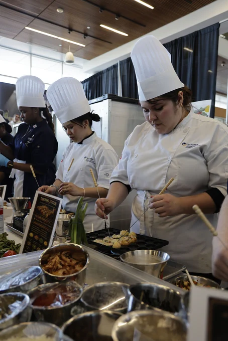 Students wearing chefs uniforms are cooking.
