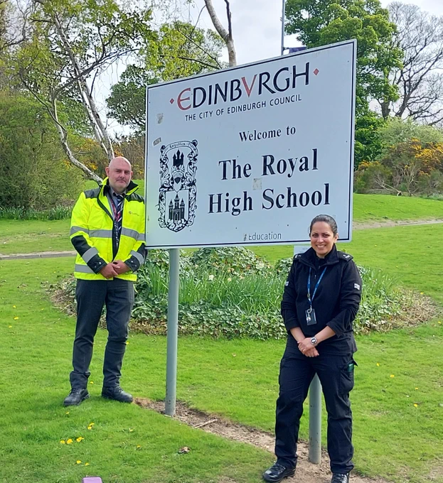 A man in a high-vis jacket and a woman in a black police uniform stand by a sign for The Royal High School in Edinburgh.