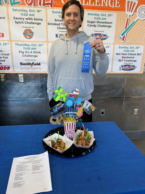 A man with brown hair in a grey sweatshirt holding a blue N.C. State Fair first-place ribbon. In front of the man is a table covered with a blue tablecloth and a tray full of fair themed food. 