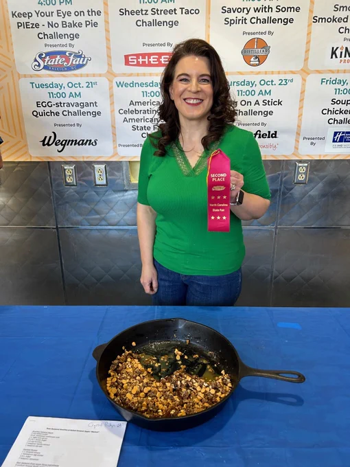 A woman with brown hair and a green top holding a red N.C. State Fair second-place ribbon. In front of the woman is a table covered in a blue tablecloth and a cast iron pan full of food on the table. 