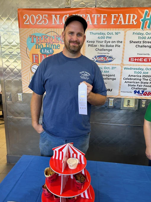 A man in a black ball cap and a blue t-shirt holding a white N.C. State Fair third-place ribbon. In front of him is a table covered in a blue tablecloth and a fair themed food item on the table. 