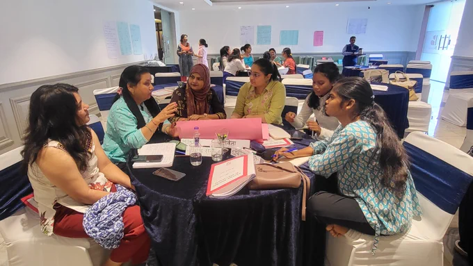 Participants sit around a round table in a workshop setting, engaged in a small-group discussion. One woman gestures as she speaks while others listen attentively, with notebooks, phones, and papers spread across the table. Additional groups and facilitators are visible in the background, and handwritten charts are displayed on the walls.

