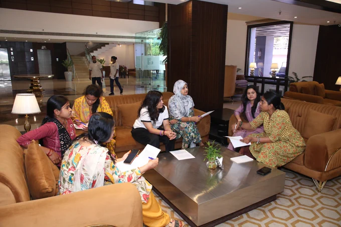 A group of women sitting on sofas in a hotel lobby, engaged in a discussion while holding papers and phones; a modern interior with a staircase and glass walls is visible in the background.
