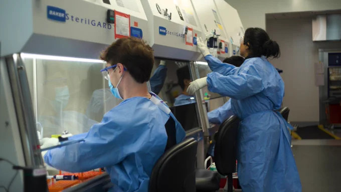 A man with reddish brown hair wears a blue surgical mask and lab goggles and looks under a lab hood to pipette liquid.