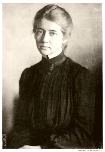 A black and white portrait of a white woman with tied-back hair, wearing a high-collared, long-sleeved dress. She gazes with a calm, neutral expression, with her hands folded in her lap.