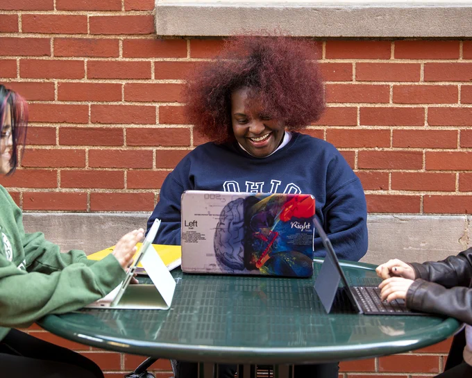 Three people are seated at an outdoor table with brick wall in the background. One person wears an "Ohio" sweatshirt and smiles at a colorful laptop. The other two have laptops and tablets.