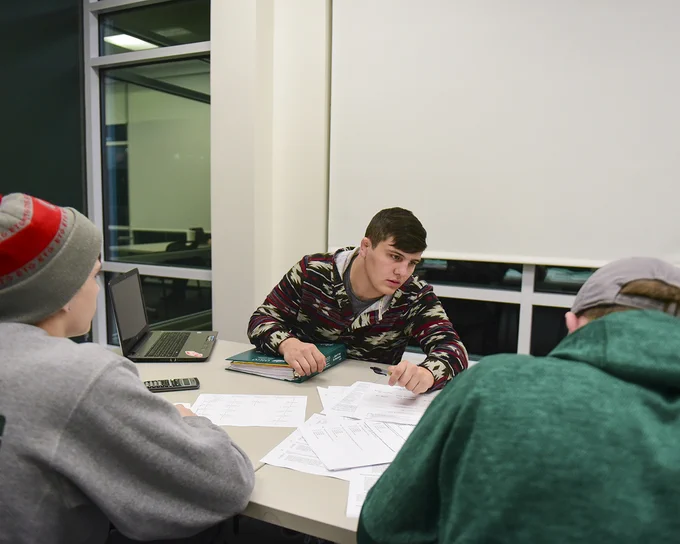 Three people are sitting at a table covered with papers in a study or meeting room. One person is speaking to the others, who are focused on the discussion. A laptop is open on the table. There is a large window in the background.