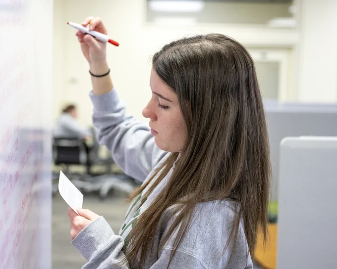 A person is writing on a whiteboard with a red marker while holding a piece of paper in the other hand. The setting appears to be an office or study space, with another individual visible in the background.