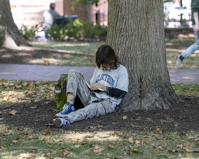 A person is sitting against a tree in a park. They are looking at their book, with a green backpack beside them. Fallen leaves are scattered on the ground.