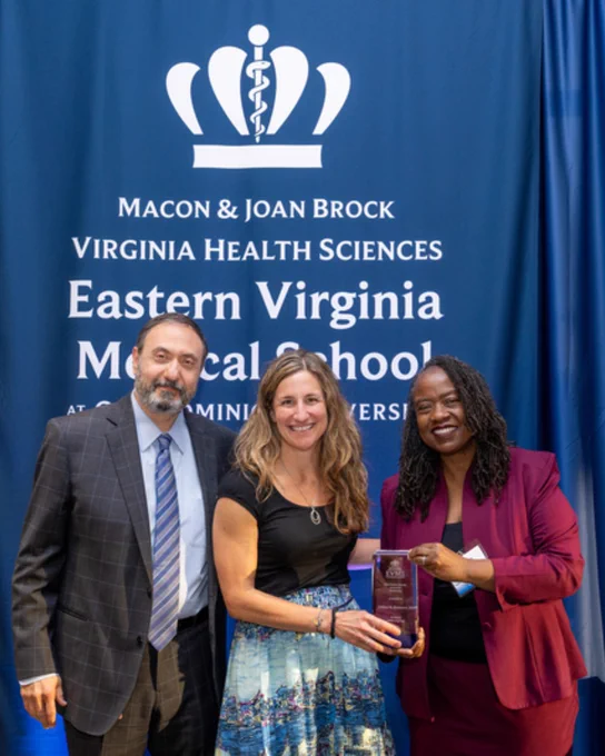 Three individuals standing in front of a blue backdrop with Eastern Virginia Medical School logo, holding an award together during a formal event.