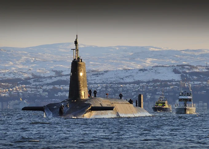 A submarine partially surfaced in calm waters with crew members visible on deck and snow-covered mountains in the background.