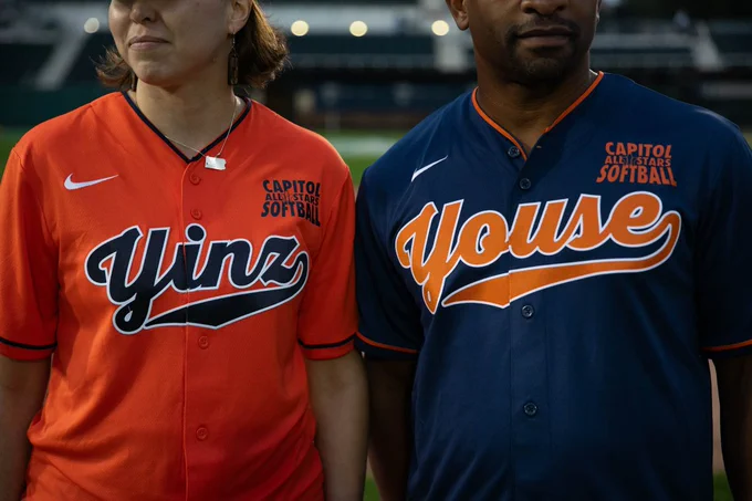 Two people in softball jerseys stand side by side; one in orange with "Yinz," the other in blue with "Youse," both labeled "Capitol All-Stars Softball."