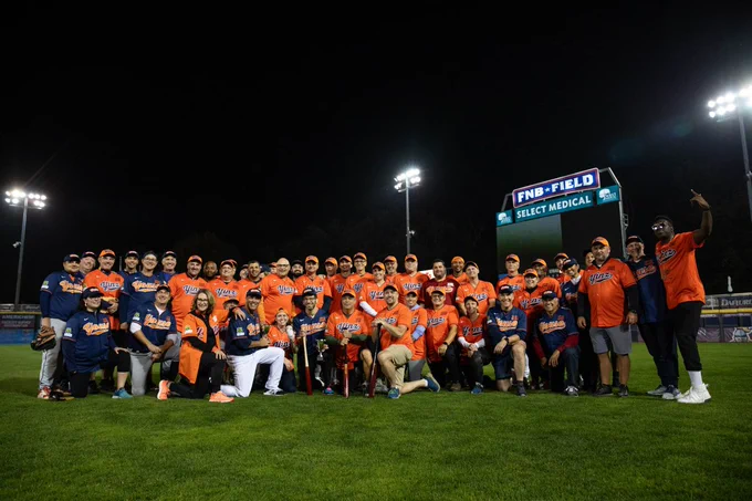 A large group of people in matching orange and navy baseball uniforms pose together on a baseball field at night, under bright stadium lights.