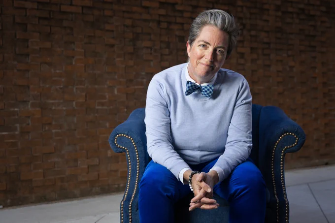 Gabrielle Calvocoressi sitting in a blue velvet chair, wearing blue pants, a blue sweater, and a blue bow tie smiling at the camera.