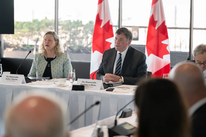 Le ministre LeBlanc prend la parole lors d’une réunion du Comité consultatif, assis à une table avec des microphones, aux côtés d’autres participants, avec des drapeaux du Canada en arrière-plan.